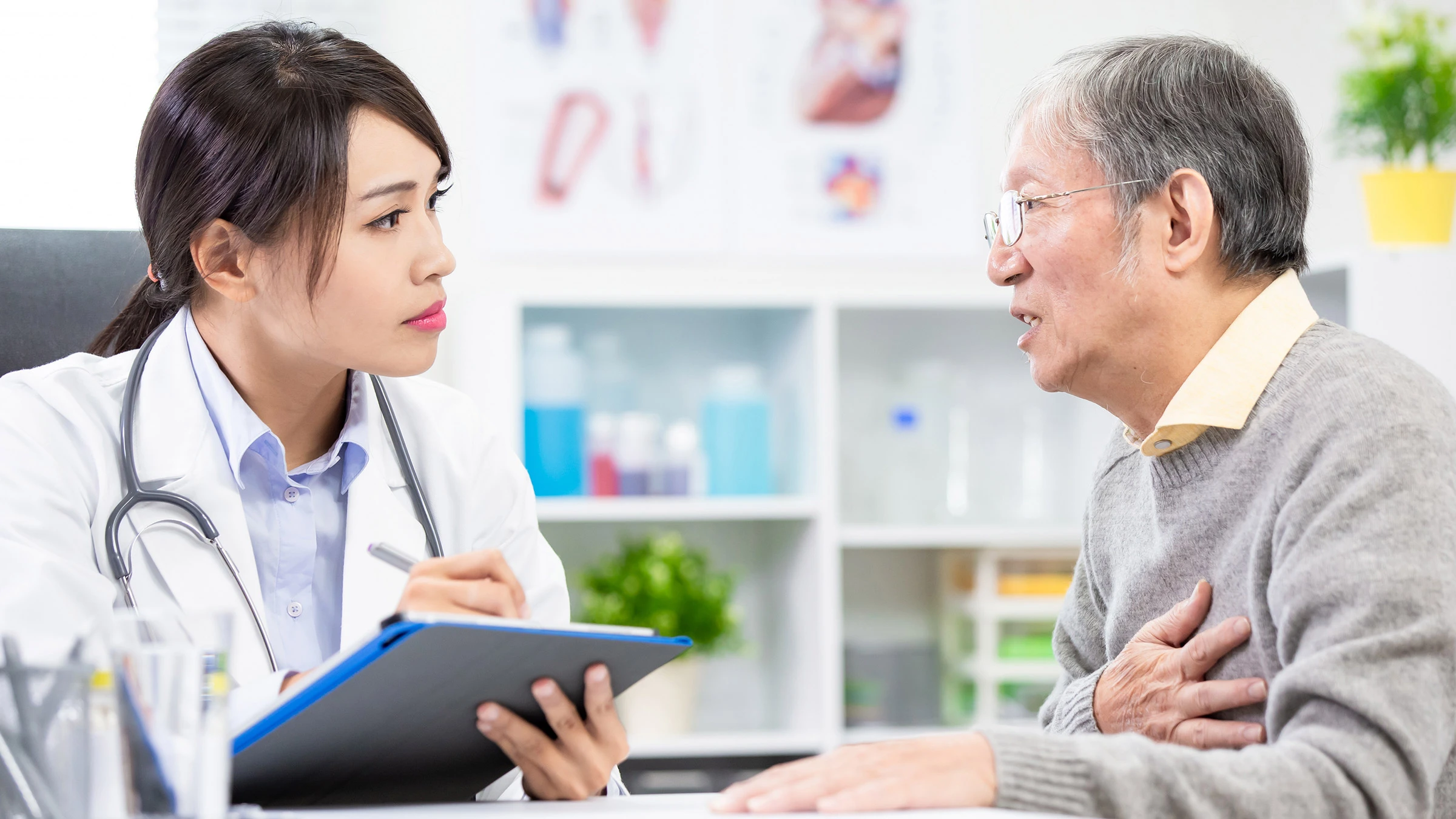 The image depicts a serious medical consultation between a young female doctor and an elderly male patient. The doctor, of Asian descent, is seated at a desk, wearing a white lab coat and stethoscope, intently focused on the patient and holding a clipboard. The elderly man is clutching his chest, appearing distressed, suggesting a potentially serious medical issue. The setting is a brightly lit doctor's office.