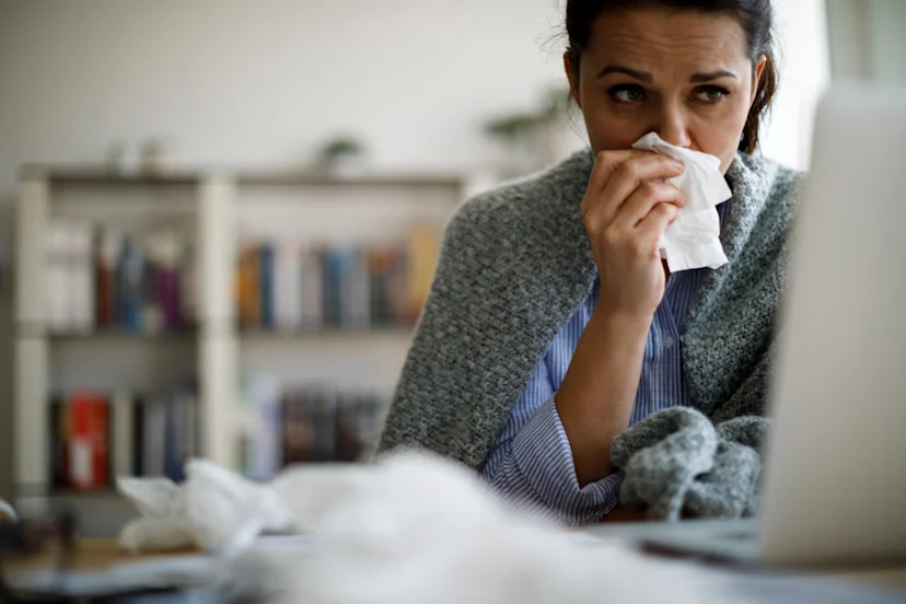 A woman bundled in a blanket looking at her computer and blowing her nose while fighting off the flu