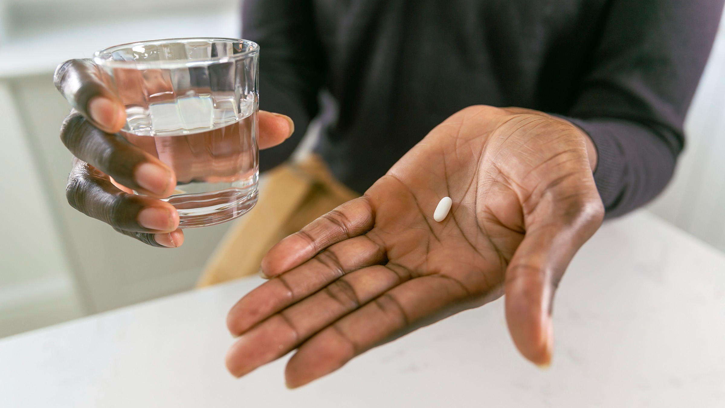 Close-up on a Black man's hand holding a single oblong pill. He is holding a glass of water in the other hand and is wearing a black shirt with tan pants.
Fly View Productions/E+ via Getty Images
