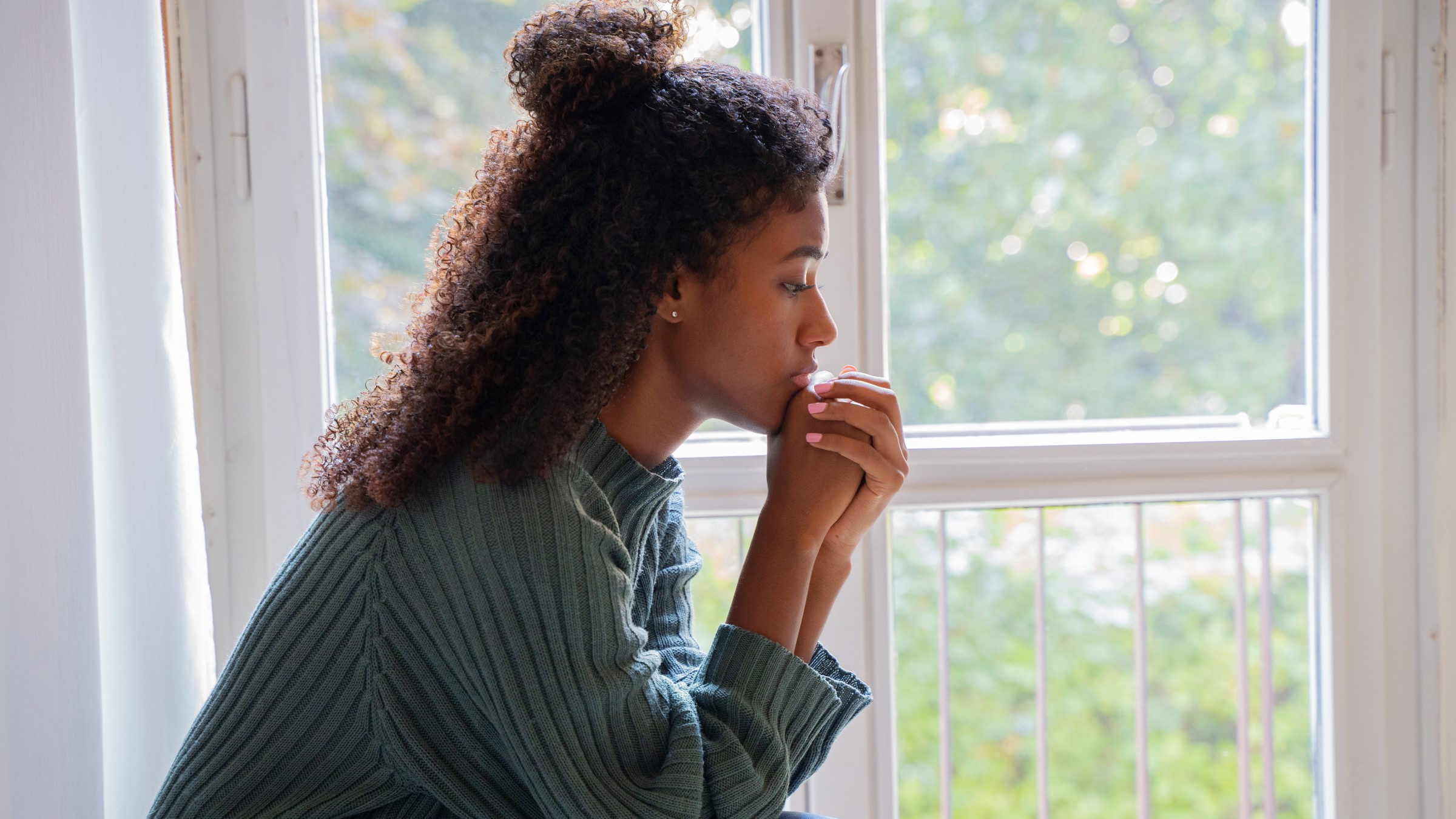 Young woman in a green sweater with curly hair sitting at a window looking depressed.
tommaso79/iStock via Getty Images