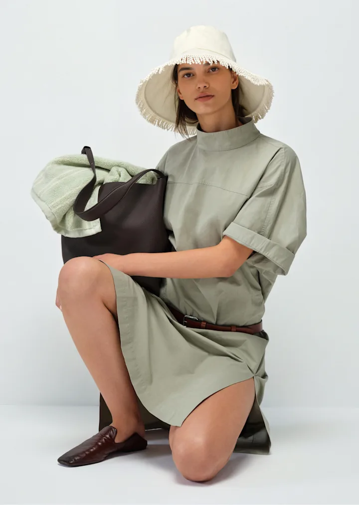Model in a sage shirt dress and canvas hat holding a leather tote