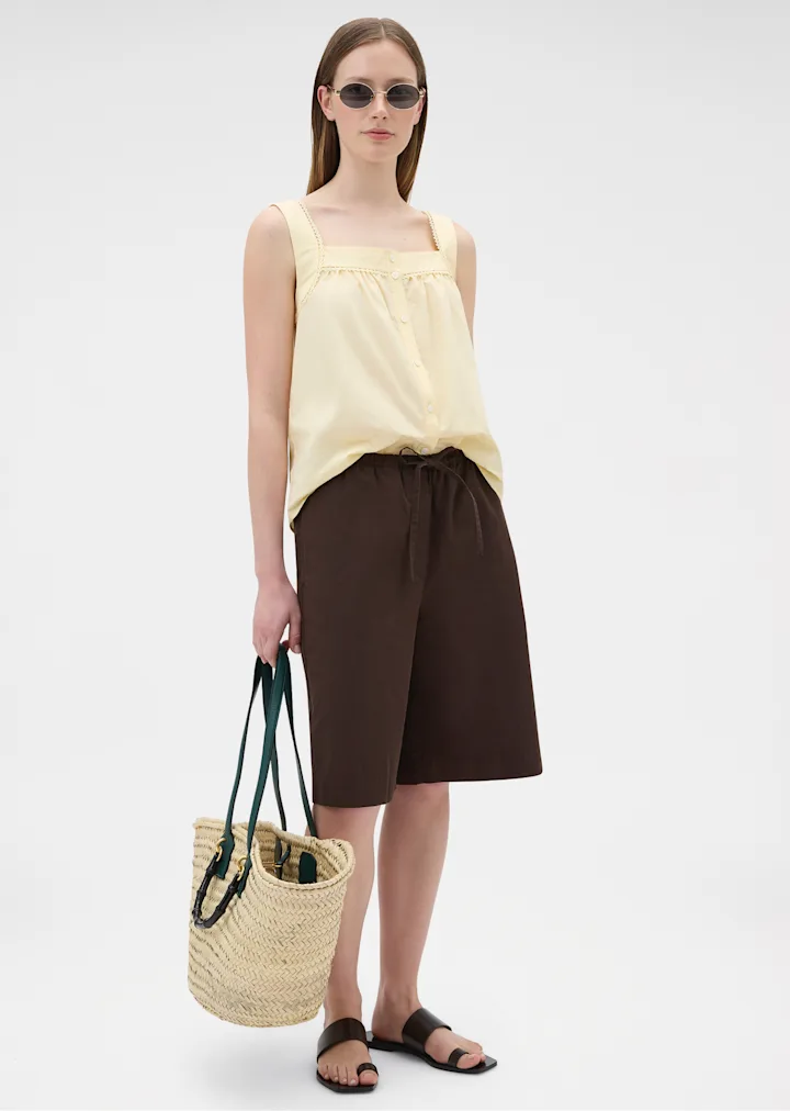 Model in a lemon camisole and brown shorts carrying a woven basket bag