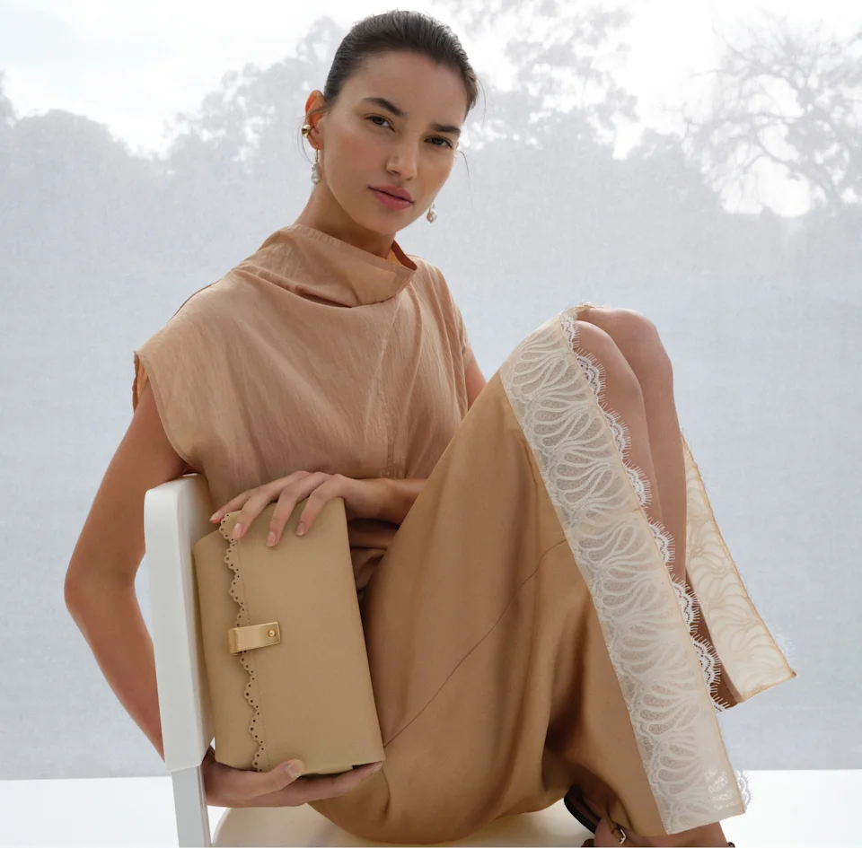 Model in a tan draped top and lace trim skirt holding a tan leather clutch on a white chair