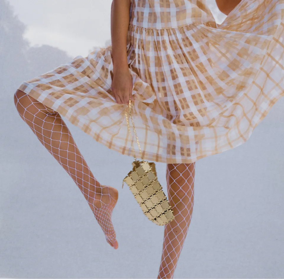 Model in a butterscotch check dress and white fishnet tights holding a gold chain bag