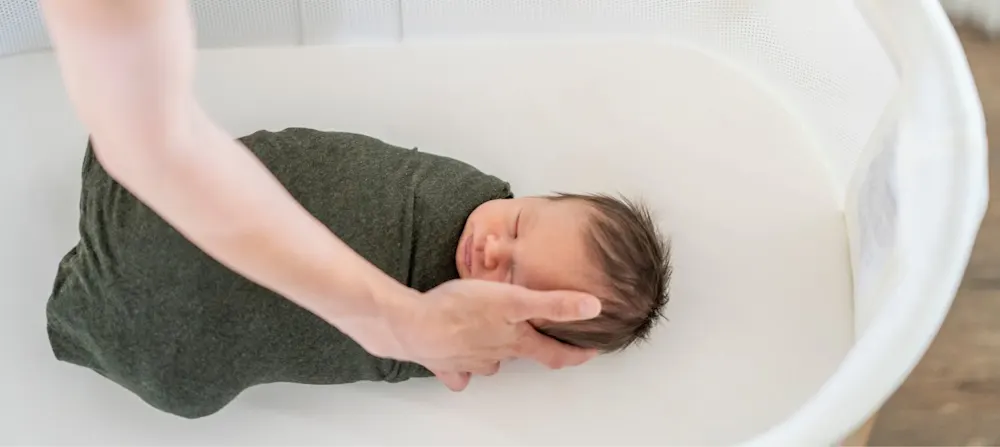 A baby is being comforted by their caregiver in a bassinet while also being swaddled.