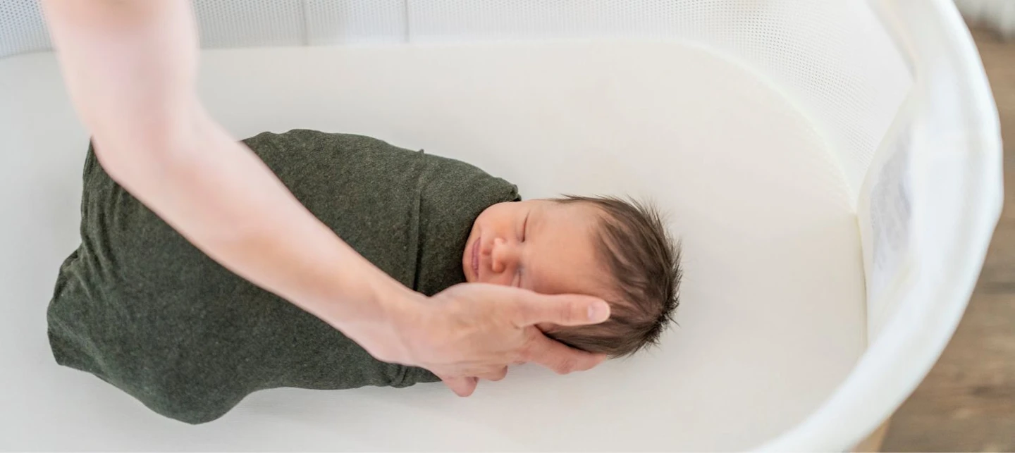 A baby is being comforted by their caregiver in a bassinet while also being swaddled. | Huckleberry