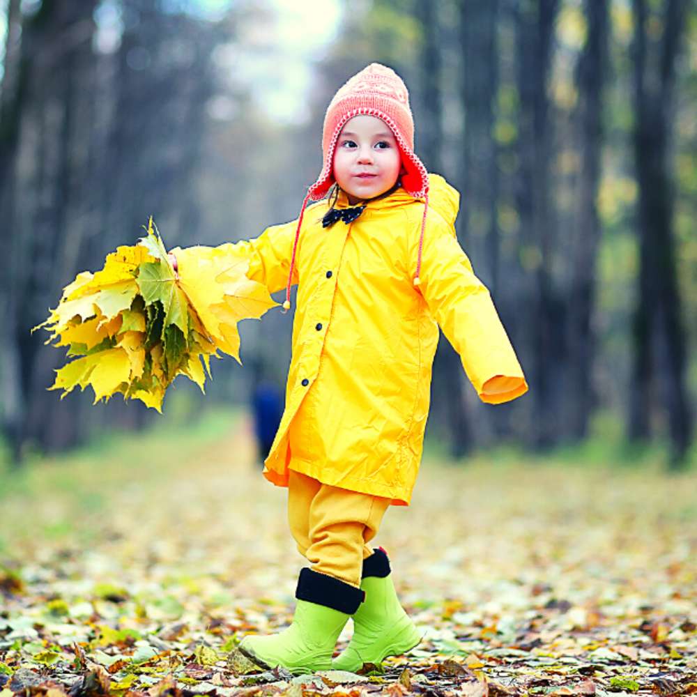 Toddler in yellow raincoat with orange beanie and green boots walking through the forest during Autumn