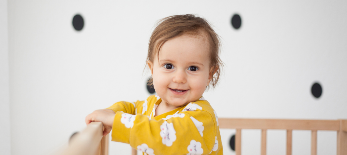 A smiling baby standing in a crib wearing a yellow shirt.