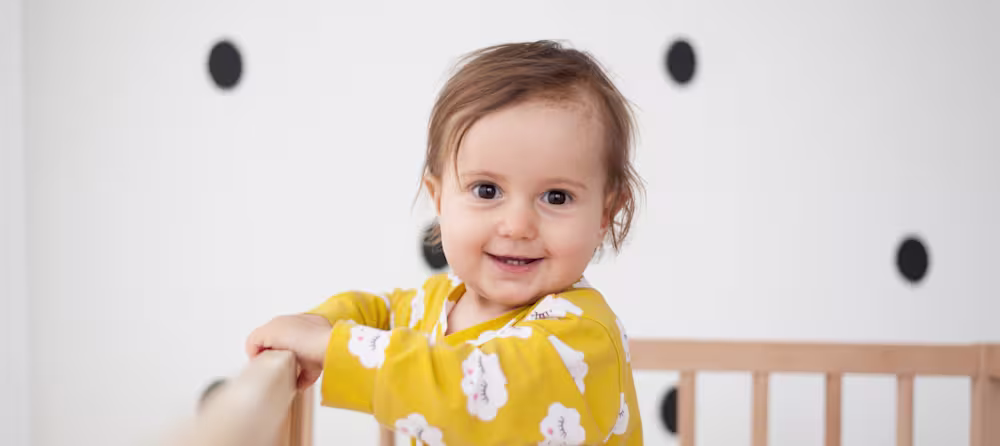 A smiling baby standing in a crib wearing a yellow shirt.
