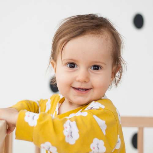 A smiling baby standing in a crib wearing a yellow shirt.