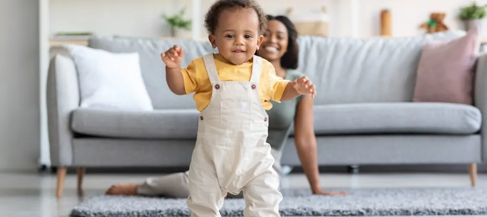 A baby starting to walk with his mother in the background.