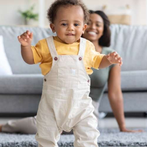 A baby starting to walk with his mother in the background.