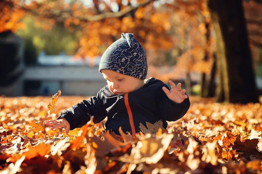 A baby sits on the floor, surrounded by leaves, happily picking them up.
