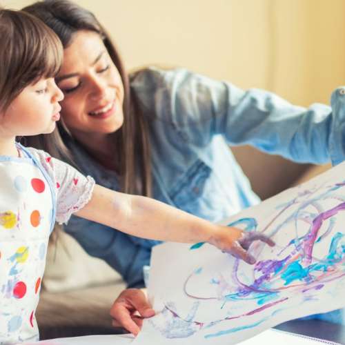 A 3 year old child finger painting with a caregiver.