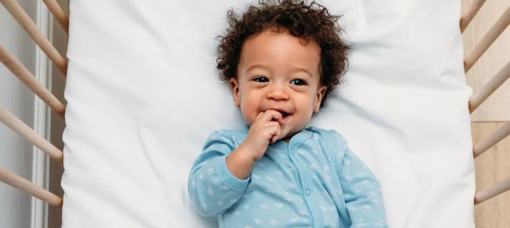 A smiling baby in a crib with a blue onesie on.