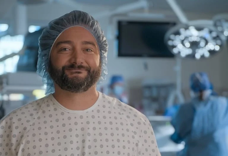 The image features a man smiling at the camera, wearing a hospital gown and a hair net.