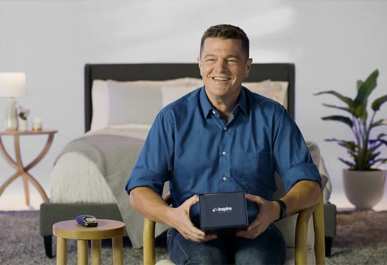 The image shows a man sitting and smiling at the camera, holding a box with the label "Inspire".