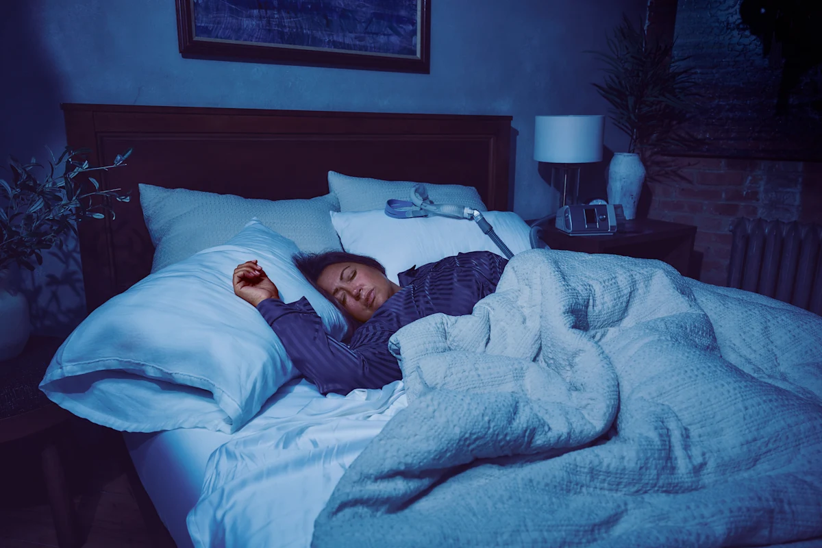 Woman lies restlessly in bed at night, with a CPAP mask and machine nearby on the nightstand.