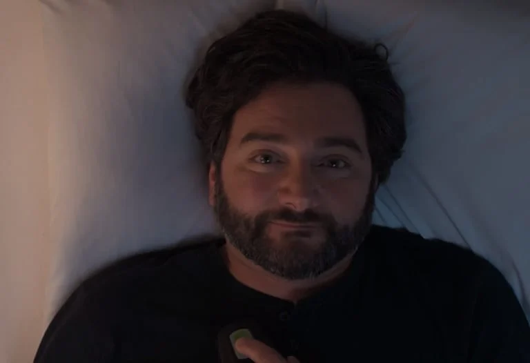 The image shows a man with a beard and dark hair, lying down with a relaxed expression.