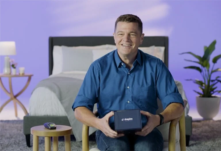 The image shows a man sitting and smiling at the camera, holding a box with the label "Inspire".