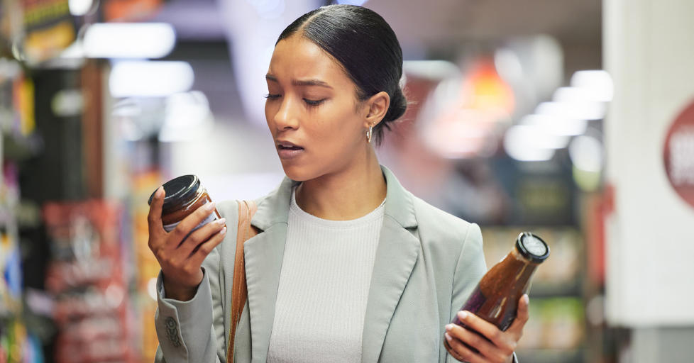 Woman wearing grey blazer and white top holding a tomato sauce in each hand reading the label one to compare the two products