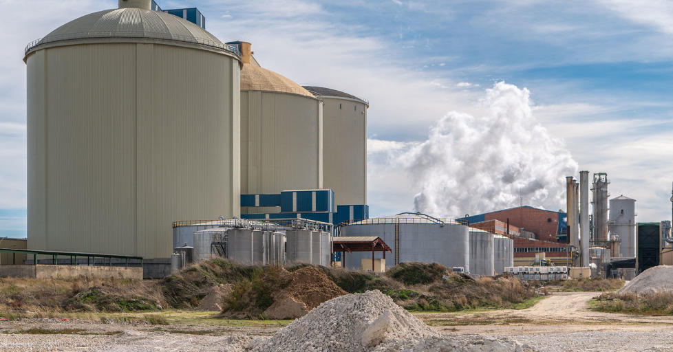 Sugar production factory with three large sugar storage buildings and chimneys in the background expelling smoke into the atmosphere