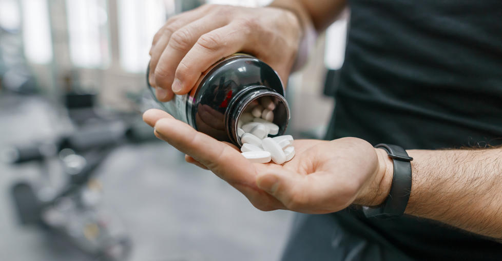 Closeup of sporty muscular man arms showing sports and fitness supplements, capsules, pills, gym background