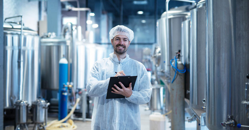 Food processing plant interior with positive smiling technologist wearing all white sanity clothing holding checklist whilst performing a check of the site