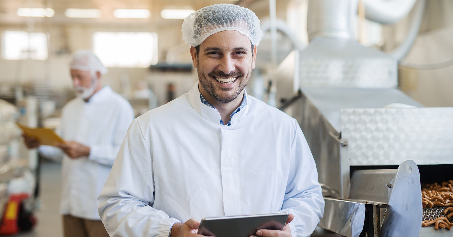 Young food technologist in a food factory holding a tablet smiles whilst walking and inspecting the factory with an older male colleague who is standing behind reviewing at a piece of paper