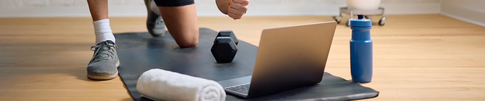 Schmuckbild: Mann, der auf einer Yoga-Matte Sport macht und ein Handtuch, eine Hantel, einen Laptop und eine Trinkflasche dabeihat.