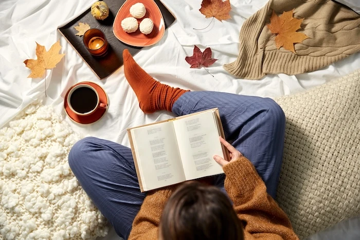 Schmuckbild: Eine Frau, die ein Buch liest, gemütliche Kleidung trägt und mit Kissen auf einem Bett sitzt. Um sie herum sind Herbstblätter, eine Tasse Kaffee, Gebäck, eine Kerze und ein Strickpullover.