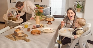 Werbung: Familie am Esstisch mit Kinderkraft Hochstuhl und Babywippe während einer gemeinsamen Mahlzeit.