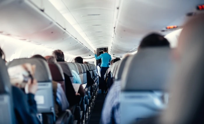 Schmuckbild: Innenansicht eines Flugzeugs mit Passagieren auf den Sitzen und einer Stewardess in Uniform, die den Gang entlanggeht und die Passagiere bedient.