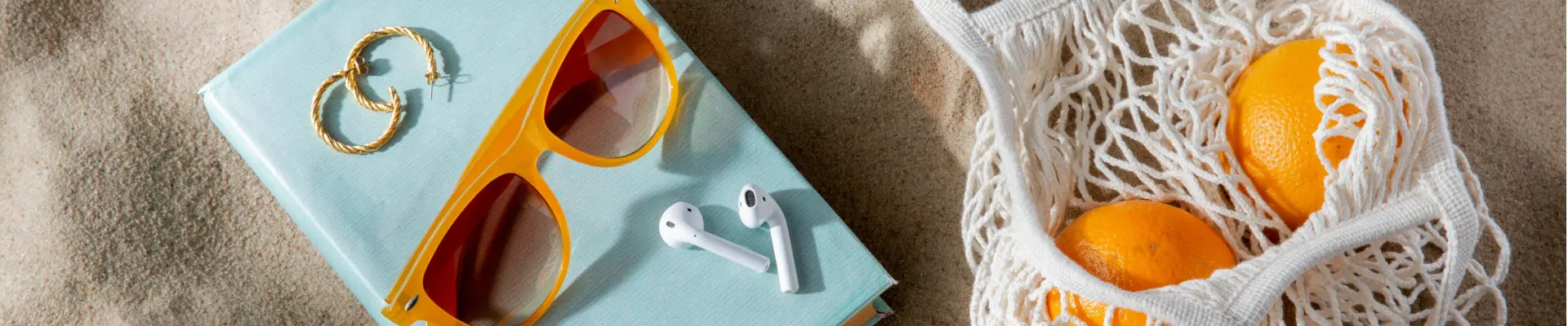 Schmuckbild: Obsttasche mit Orangen, Buch, Sonnenbrille, Ohrringe und Kopfhörer auf Sand