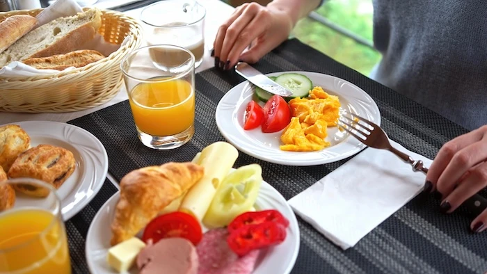 Schmuckbild: Ein Frühstückstisch mit verschiedenen Tellern, Gläsern und Tassen sowie einem Brotkorb. Eine Person hält ein Messer und eine Gabel, auf den Tellern sind Speisen wie Rührei, Croissants, Wurst und Käse.