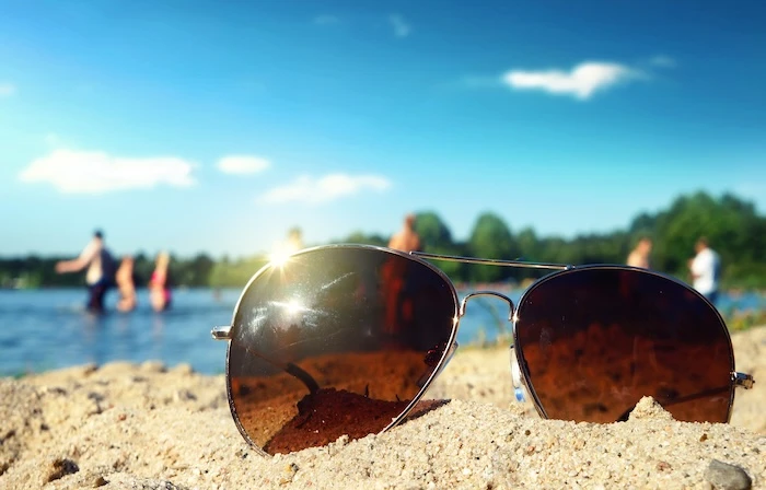 Schmuckbild: Eine Sonnenbrille im Sand, dahinter ein Badesee mit Menschen im Wasser.