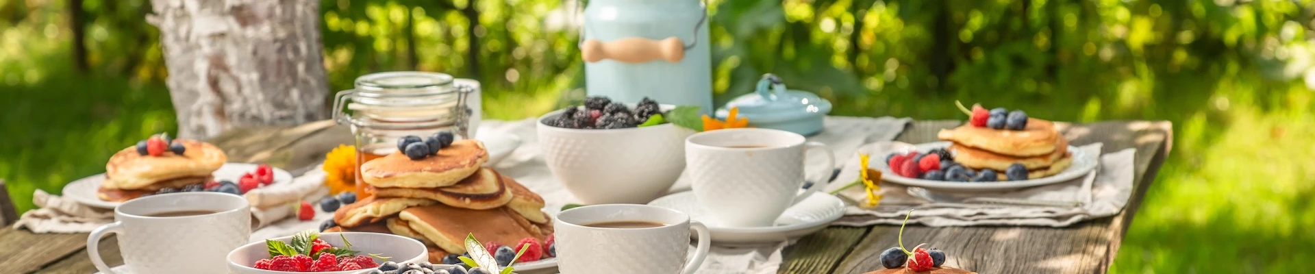 Schmuckbild: Frühstückstisch im Garten, darauf Besteck, Geschirr wie Tassen und Teller, gefüllt mit Pancakes und Früchten.