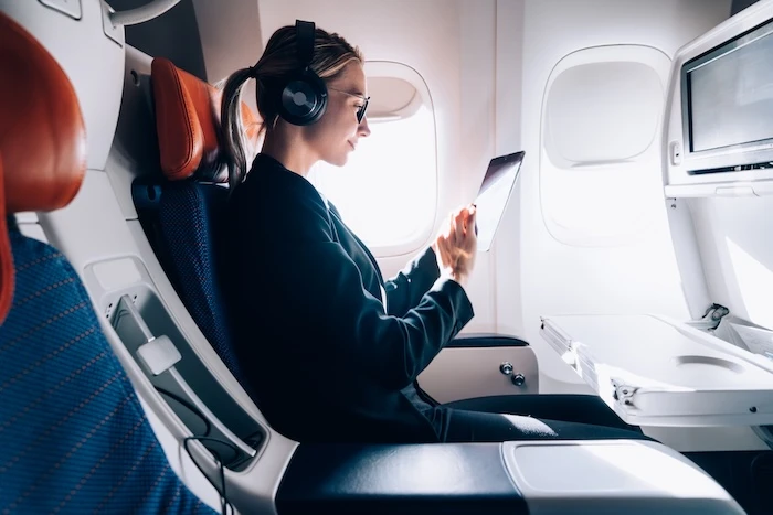 Schmuckbild: Frau, die mit Kopfhörern und einem Tablet in der Hand in einem Flugzeugsitz sitzt.