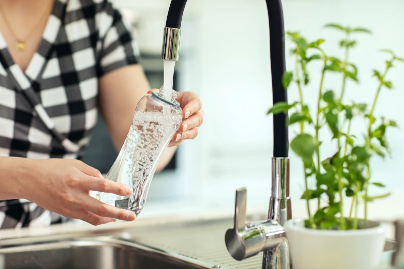 Schmuckbild: Person hält Glasflasche unter einen Wasserhahn und befüllt sie mit Wasser. Neben dem Wasserhahn ist eine Basilikumpflanze in einem Topf zu sehen.