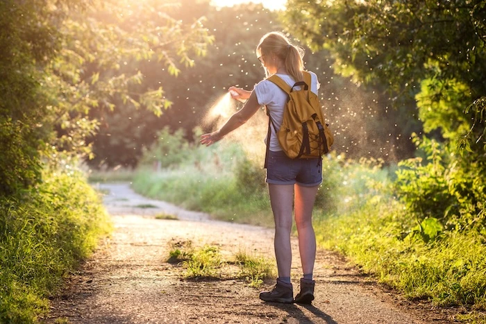 Schmuckbild: Eine Frau mit Rucksack und Wanderschuhen auf einem Waldweg, ringsherum Gebüsch und zahlreiche Stechmücken. Sie hält eine Sprühflasche in der Hand und sprüht sich ein.