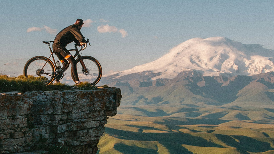 Werbung: Ein Radfahrer auf einem Felsvorsprung blickt auf ein beeindruckendes, schneebedecktes Bergpanorama.
