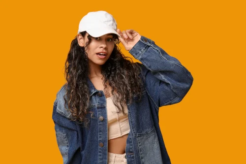 Schmuckbild: Eine Frau trägt ein weißes Cap sowie eine Jeansjacke und ein kurzes Top und fasst mit einer Hand an den Schirm der Kappe