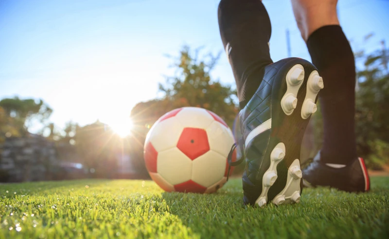 Schmuckbild: Grüner Rasen, auf dem ein rot-weißer Fußball liegt. Dahinter steht ein Mensch mit Fußballschuhen, der den Fuß anwinkelt.