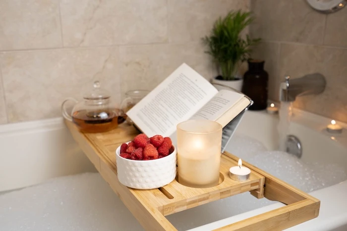 Badewannenablage auf Badewanne, darauf eine schale Erdbeeren, Tee, ein Buch und Kerzen
