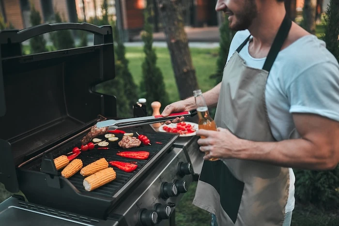 Schmuckbild: Mann, der mit einer Schürze und einem Bier in der Hand am Grill steht.
