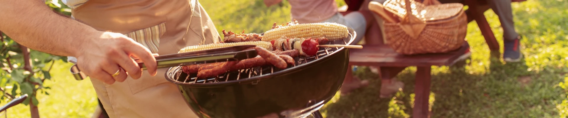 Schmuckbild: Mann, der an einem Kugelgrill steht und verschiedene Lebensmittel grillt. Im Hintergrund Wiese und ein Picknicktisch, an dem Personen sitzen.