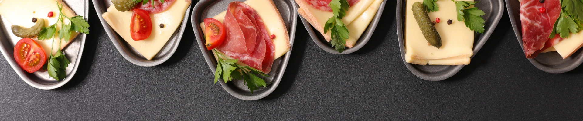 Schmuckbild: Mehrere Raclette-Pfännchen nebeneinander mit Käse und Wurst befüllt