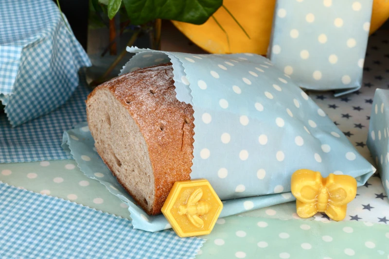 Schmuckbild: Ein angeschnittenes Laib Brot, das halb in ein Bienenwachstuch eingewickelt ist. Davor stehen Wachsfiguren eines Schmetterlings und einer Biene in einer Wabe.