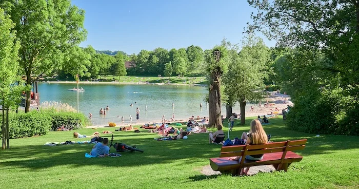 Schmuckbild: Ein Badesee mit Strandabschnitt du grüner Wiese. Mehrere Menschen Baden und entspannen am See.