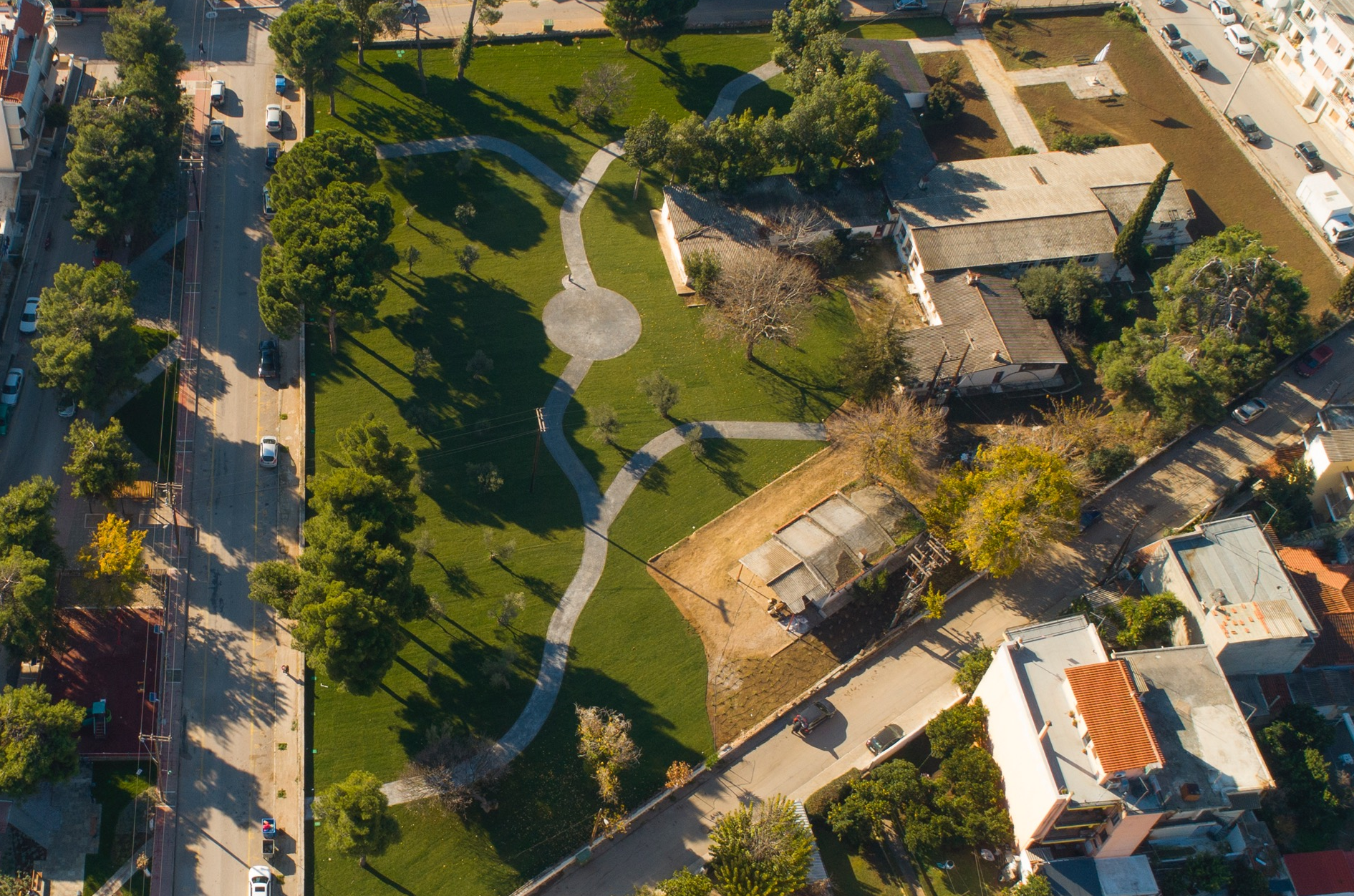 Ένα τοπίο που απεικονίζει μια περιοχή πάρκου με γρασίδι, δέντρα, μονοπάτια με στροφές και μια κεντρική κυκλική πλακόστρωτη περιοχή, που περιβάλλεται από κτίρια και δρόμους.